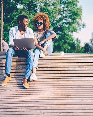 Two smiling friends outside sitting on bench looking at computer